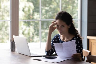 Woman looking over paperwork carefully with her laptop open.