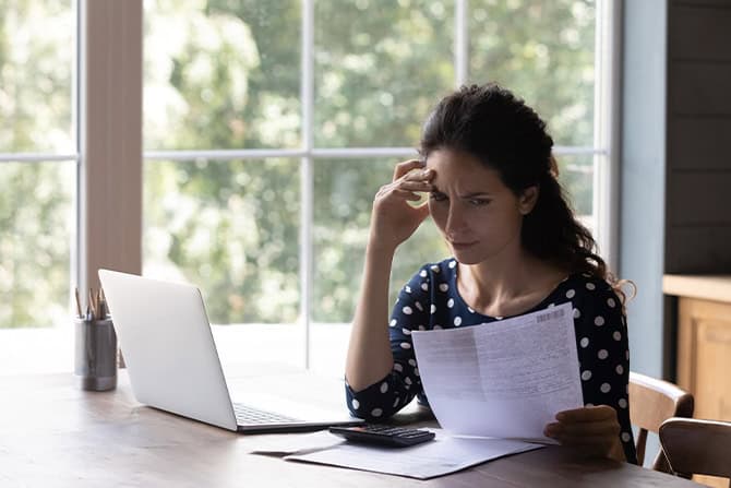 Woman looking over paperwork carefully with her laptop open.
