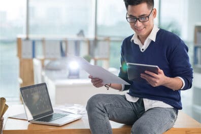 Man sitting on the edge of his desk looking at an ipad and papers to review his accounts.
