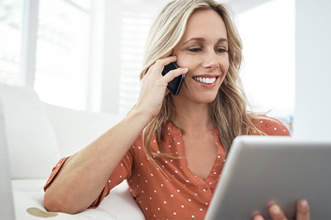 Women sitting on her couch talking on the phone while looking at her laptop.